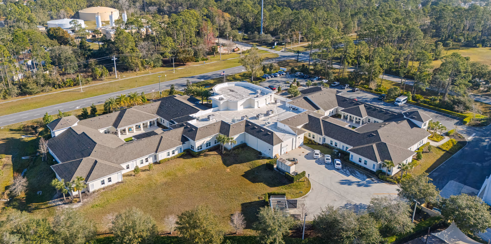 Aerial view of a large senior living facility named Artis of Palm Coast surrounded by trees and greenery, with multiple connected buildings, parking areas with cars, and a road nearby.