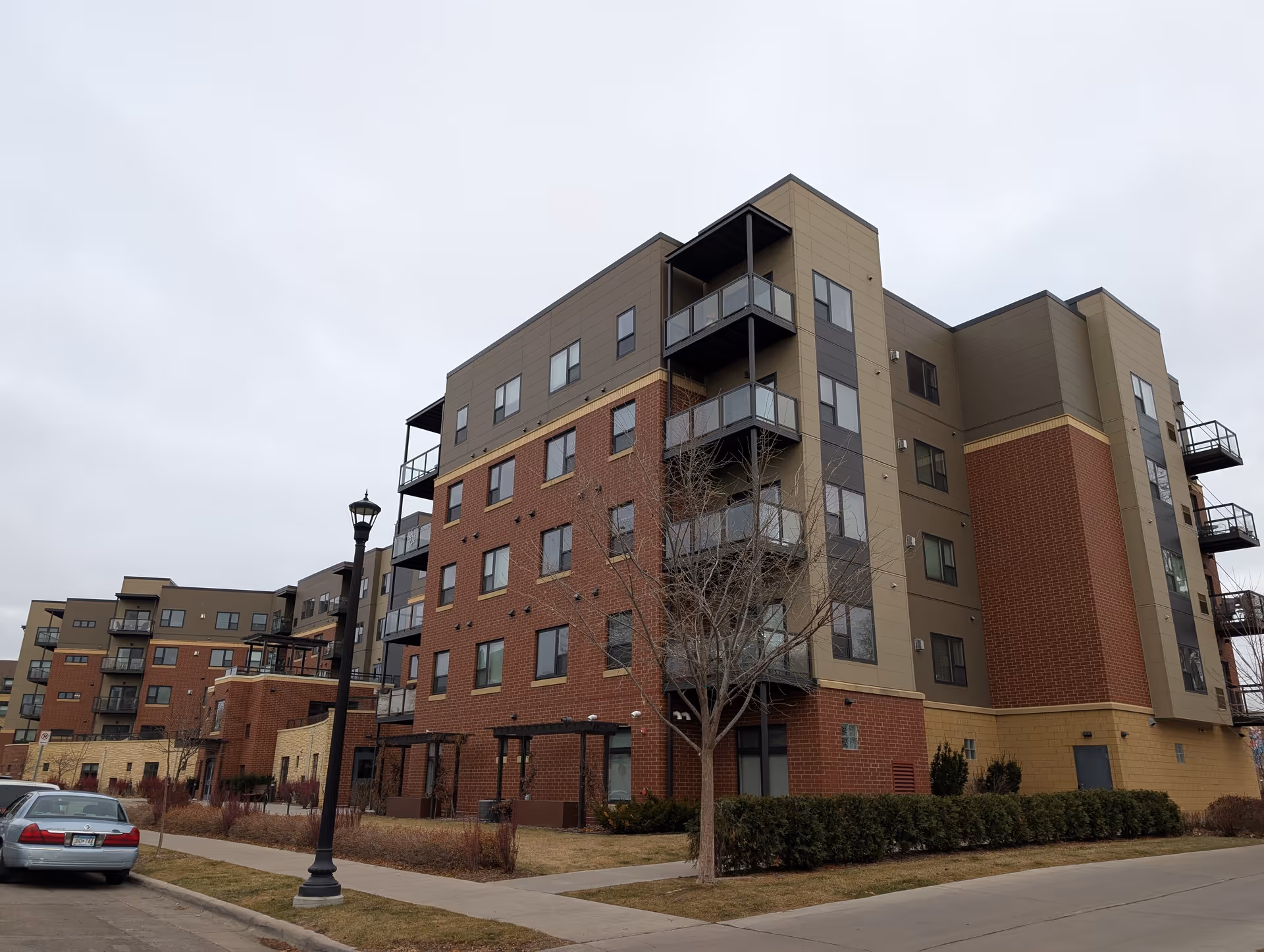 Multi-story brick and siding senior living apartment building with balconies, a streetlamp, and parked cars under an overcast sky.