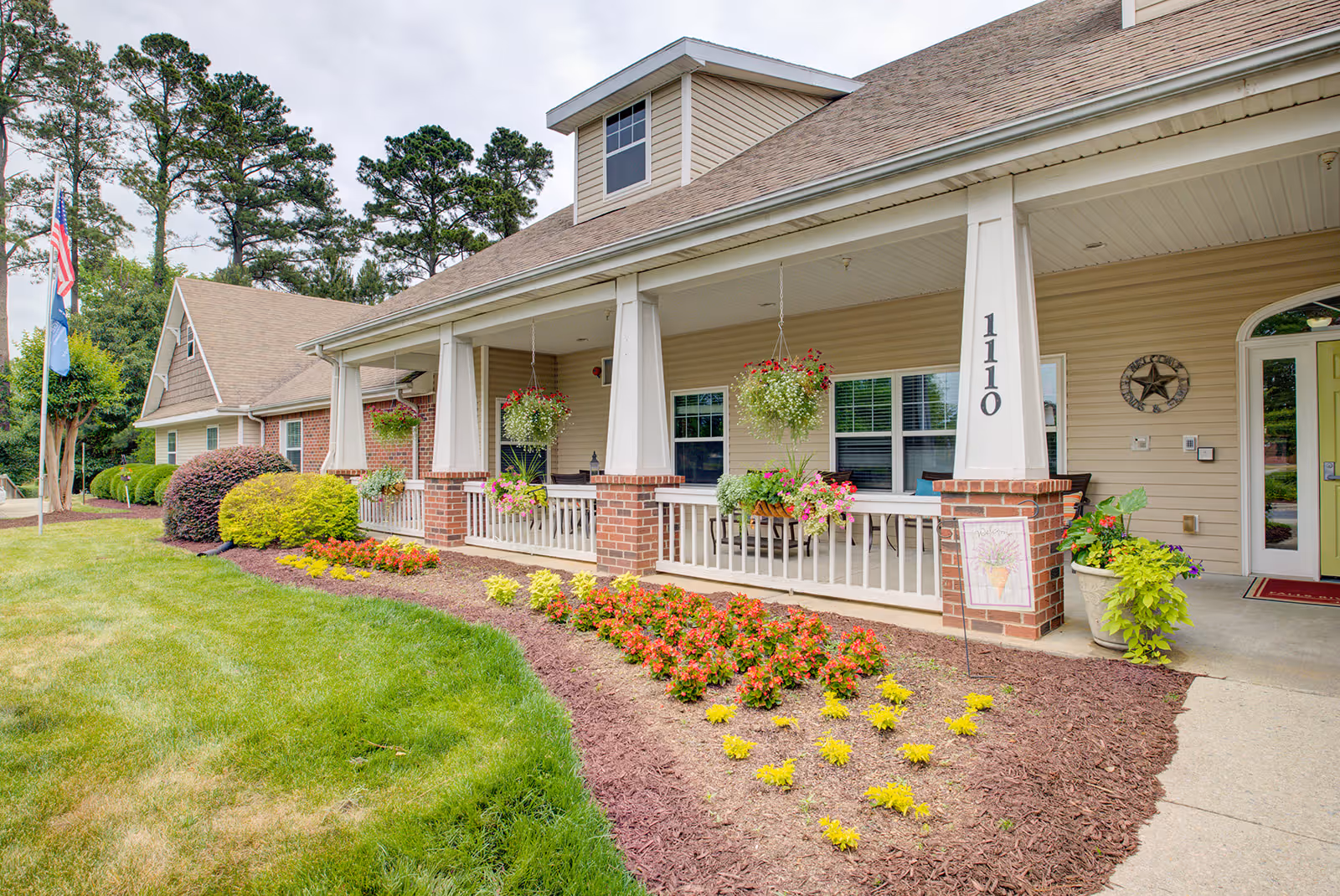 Exterior view of a single-story assisted living facility building with beige siding and brick accents. The building has a covered porch with hanging flower baskets and potted plants. A well-maintained garden with colorful flowers and green shrubs lines the walkway leading to the entrance. The building number 1110 is visible on a porch column, and flags are seen in the background near tall trees.