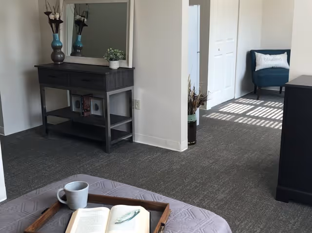 A cozy interior space featuring a gray carpeted floor, a dark wooden console table with decorative vases and a small plant, a large mirror above the table, and a blue armchair with a pillow that says 'blessed' near a white door. In the foreground, there is a tray with a white mug and an open book resting on a gray quilted surface.