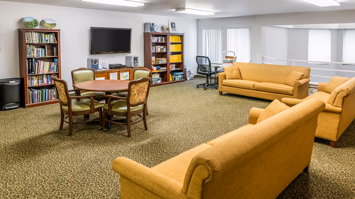 A cozy common area in a senior living facility with three mustard-yellow sofas arranged around a patterned carpet. There is a round wooden table with four chairs, two wooden bookshelves filled with books and magazines, a flat-screen TV mounted on the wall, and a desk with an office chair near windows with vertical blinds.