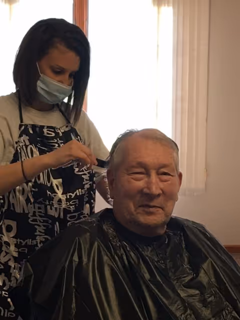 A woman wearing a face mask and an apron is cutting the hair of an elderly man who is seated and covered with a black haircut cape in a well-lit room with curtains.
