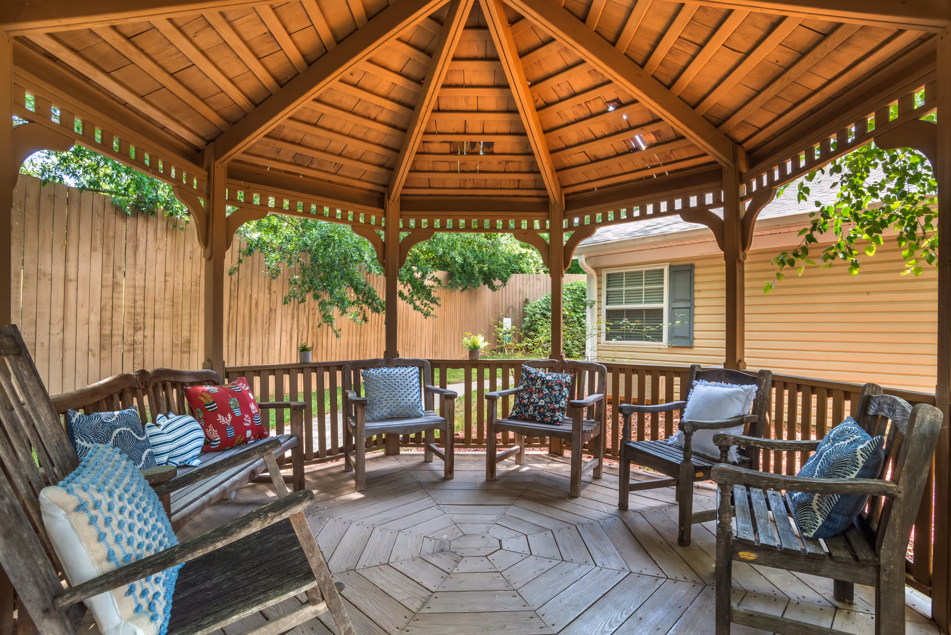 Wooden gazebo with cushioned wooden chairs arranged around its center in a fenced backyard beside a house.