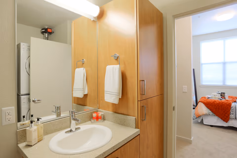 Bathroom vanity area with a sink, countertop, soap dispensers, and a large mirror. A white towel hangs on a towel ring attached to a wooden cabinet. In the background, a partially open door reveals a stacked washer and dryer. Beyond the bathroom, a bedroom with a bed covered in white and orange bedding is visible through an open doorway.