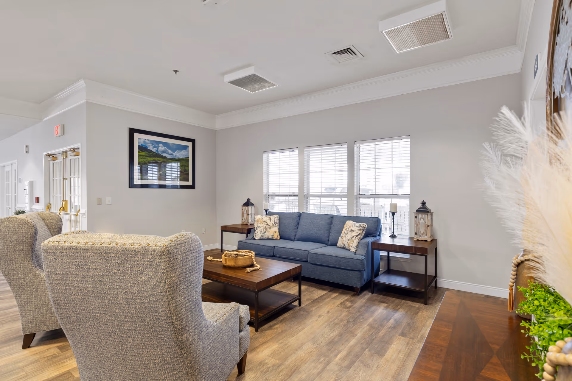 A bright and cozy living room area with a blue sofa adorned with floral pillows, two gray upholstered armchairs, a wooden coffee table, and two matching wooden side tables with decorative lanterns and candles. The room has large windows with white blinds, light-colored walls, and wood flooring. A framed landscape photograph hangs on the wall, and there is a decorative plant and pampas grass on the right side.