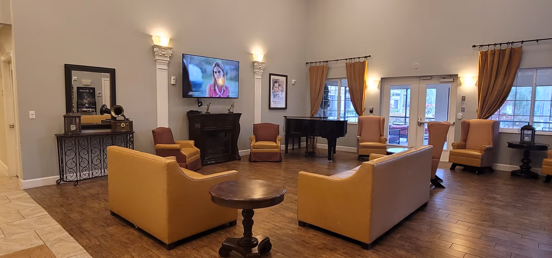 A spacious living room area with wooden flooring, featuring two mustard yellow sofas facing each other with a round wooden table in between. There are several armchairs in mustard and red colors arranged around the room. A flat-screen TV is mounted on the wall above a dark wooden fireplace. A black grand piano is placed near the windows with mustard curtains. The room is softly lit with wall sconces and has large windows and a glass door leading outside.