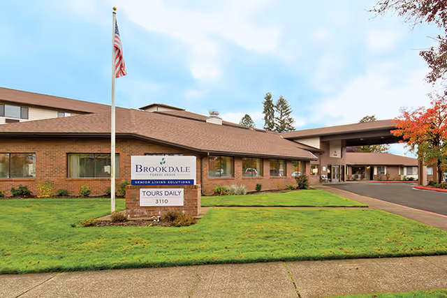 Front exterior of Brookdale Forest Grove senior living facility showing the building, entrance canopy, flagpole, and a lawn with a 'Brookdale' sign.