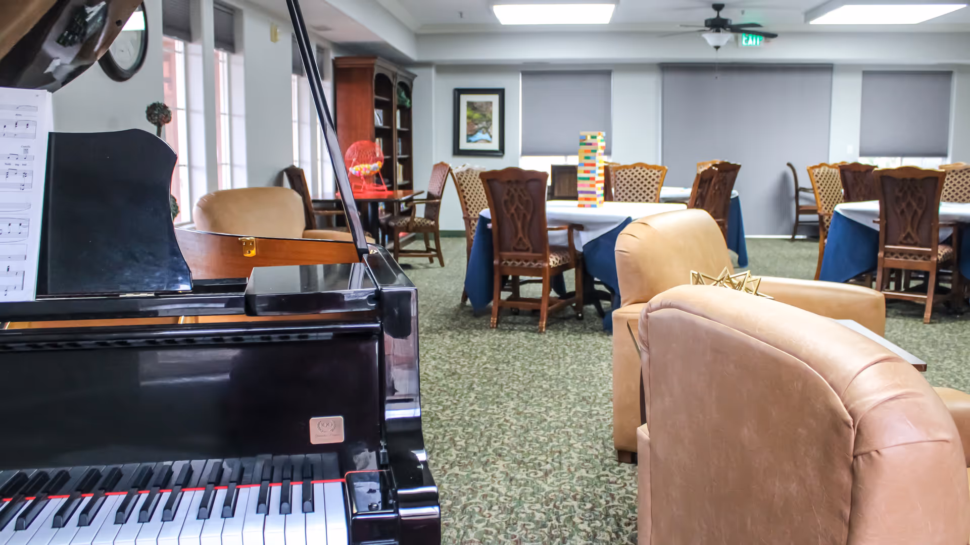 Interior view of a senior living facility common area featuring a black grand piano with sheet music, several tables with chairs, a bookshelf, and comfortable tan armchairs. The room has carpeted flooring and windows with blinds.