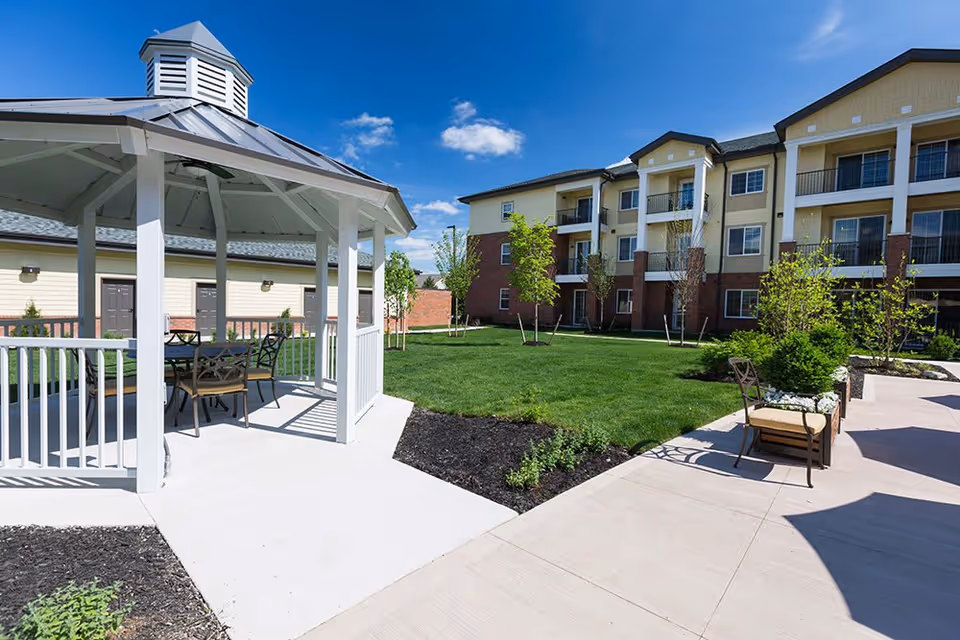 Outdoor courtyard area of a senior living facility featuring a white gazebo with a table and chairs underneath, surrounded by green grass, small trees, and a three-story building with balconies in the background under a clear blue sky.