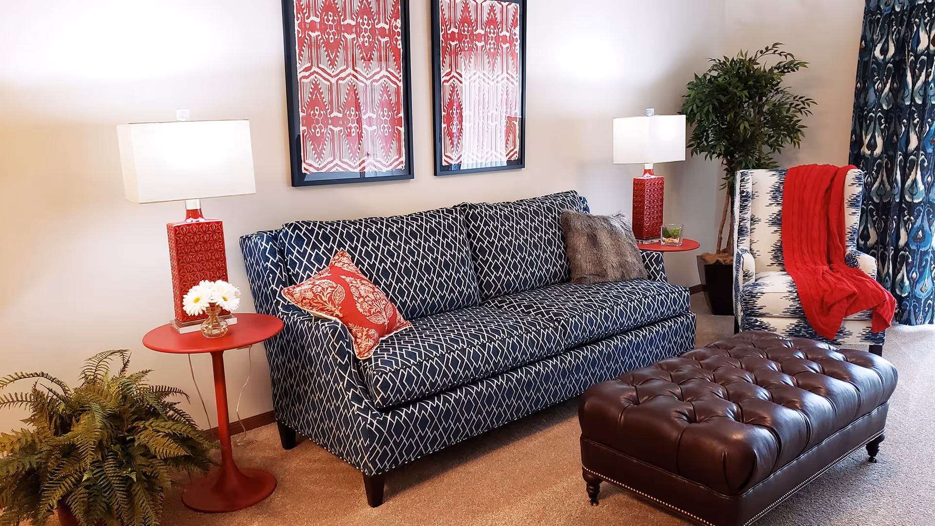 A cozy living room featuring a blue patterned sofa with red and gray pillows, a matching armchair with a red throw blanket, two red side tables with white lamps, a large tufted brown leather ottoman, two red and white framed artworks on the wall, a green potted plant, and blue patterned curtains.
