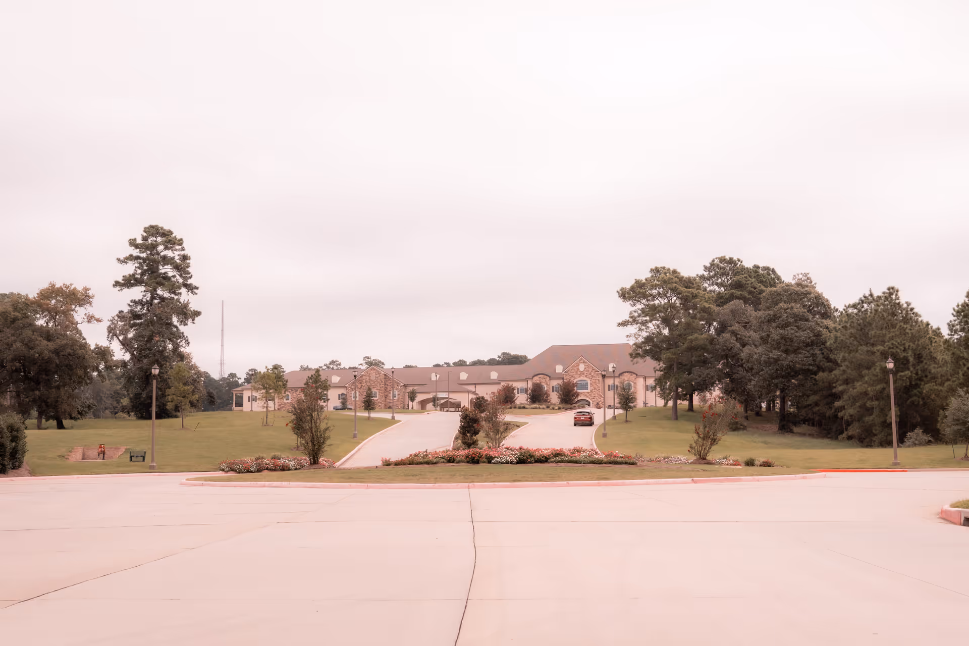 Wide view of Worthington Manor building from the parking lot, showing a long driveway leading up to the entrance surrounded by green lawns, trees, and flower beds under a cloudy sky.