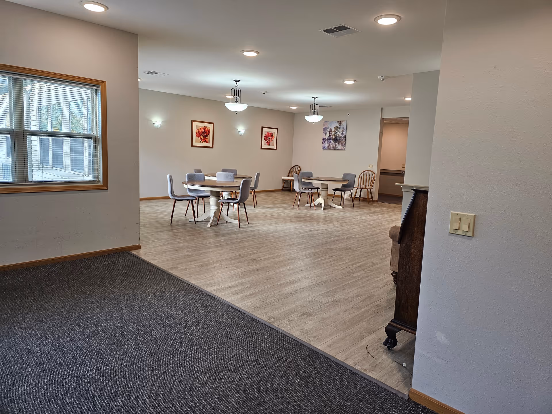 Interior view of a senior living facility common area with round tables and chairs arranged on a wood floor. The walls are decorated with framed floral and nature artwork. There are ceiling lights and wall sconces providing illumination. A window with blinds is visible on the left side.