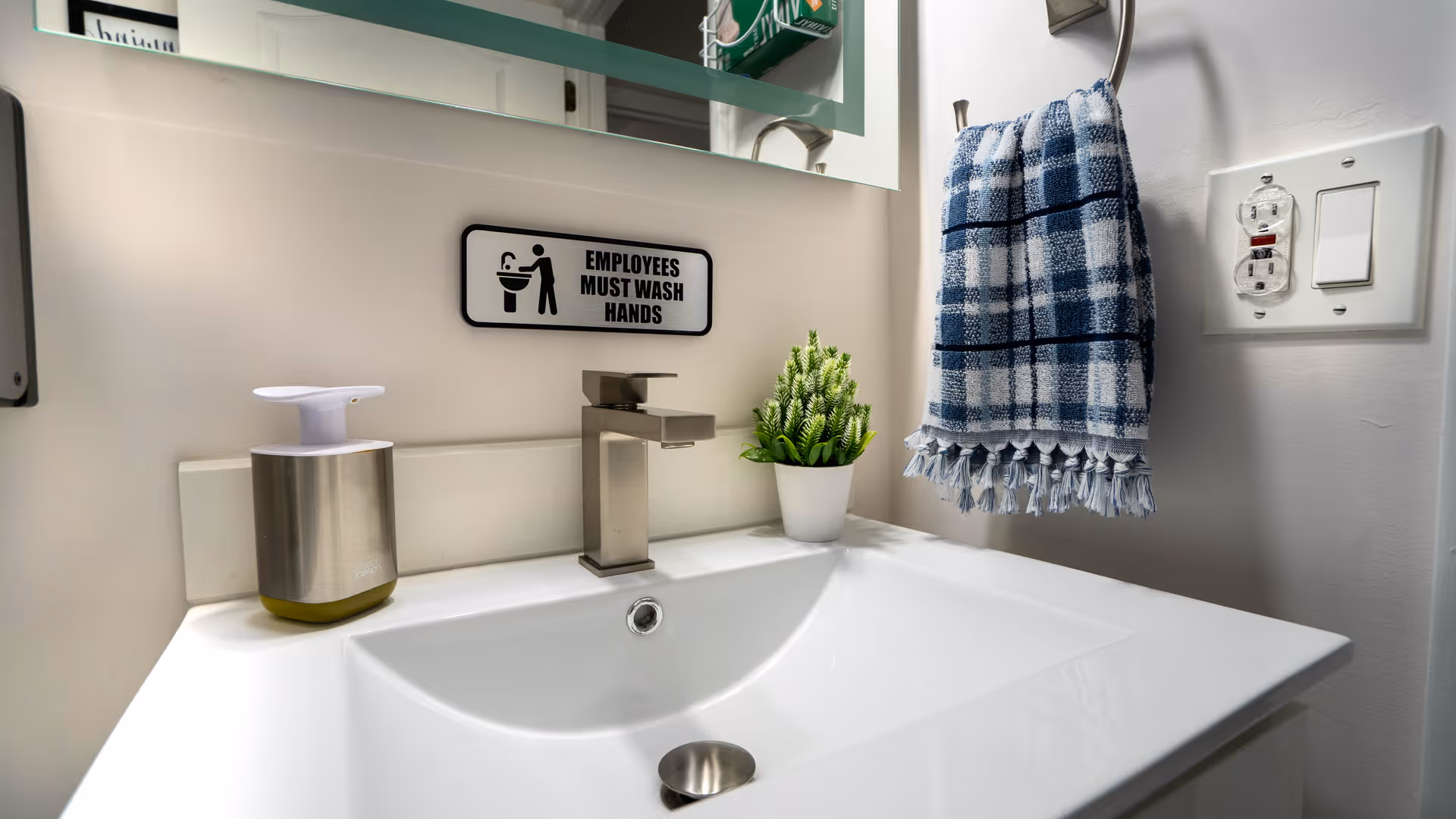 A clean bathroom sink area with a modern faucet, a soap dispenser, a small potted plant, a blue and white checkered hand towel hanging on a wall hook, and a sign above the sink that reads 'EMPLOYEES MUST WASH HANDS'.