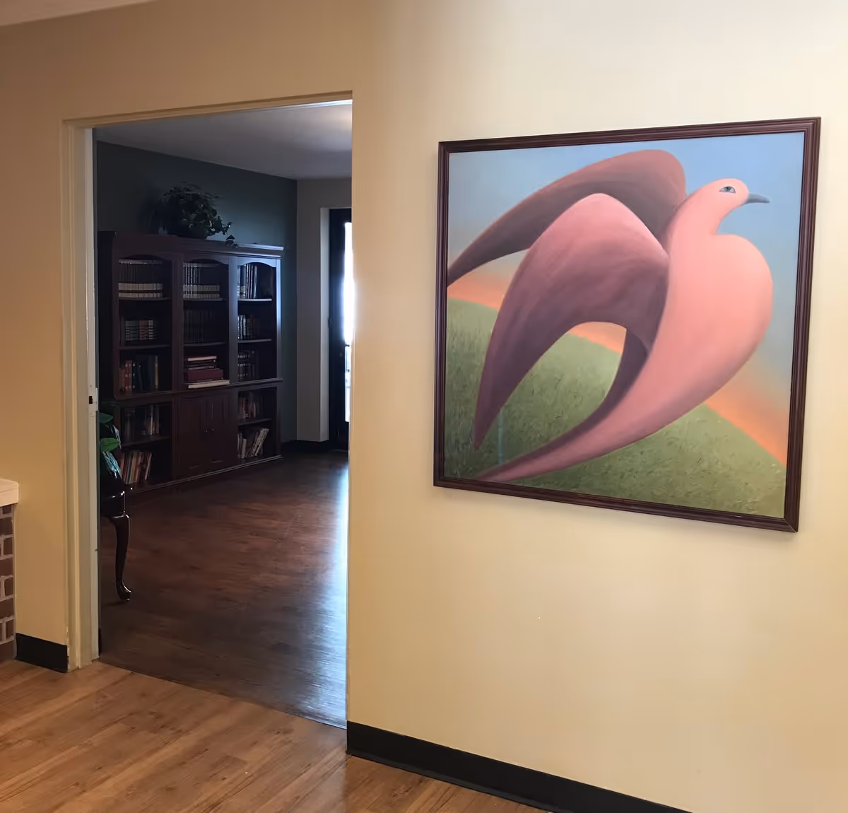 Interior view of a memory care residence showing a hallway with wooden flooring and a large framed painting of a pink bird on the wall. Through an open doorway, a room with a wooden bookshelf filled with books and a plant on top is visible.