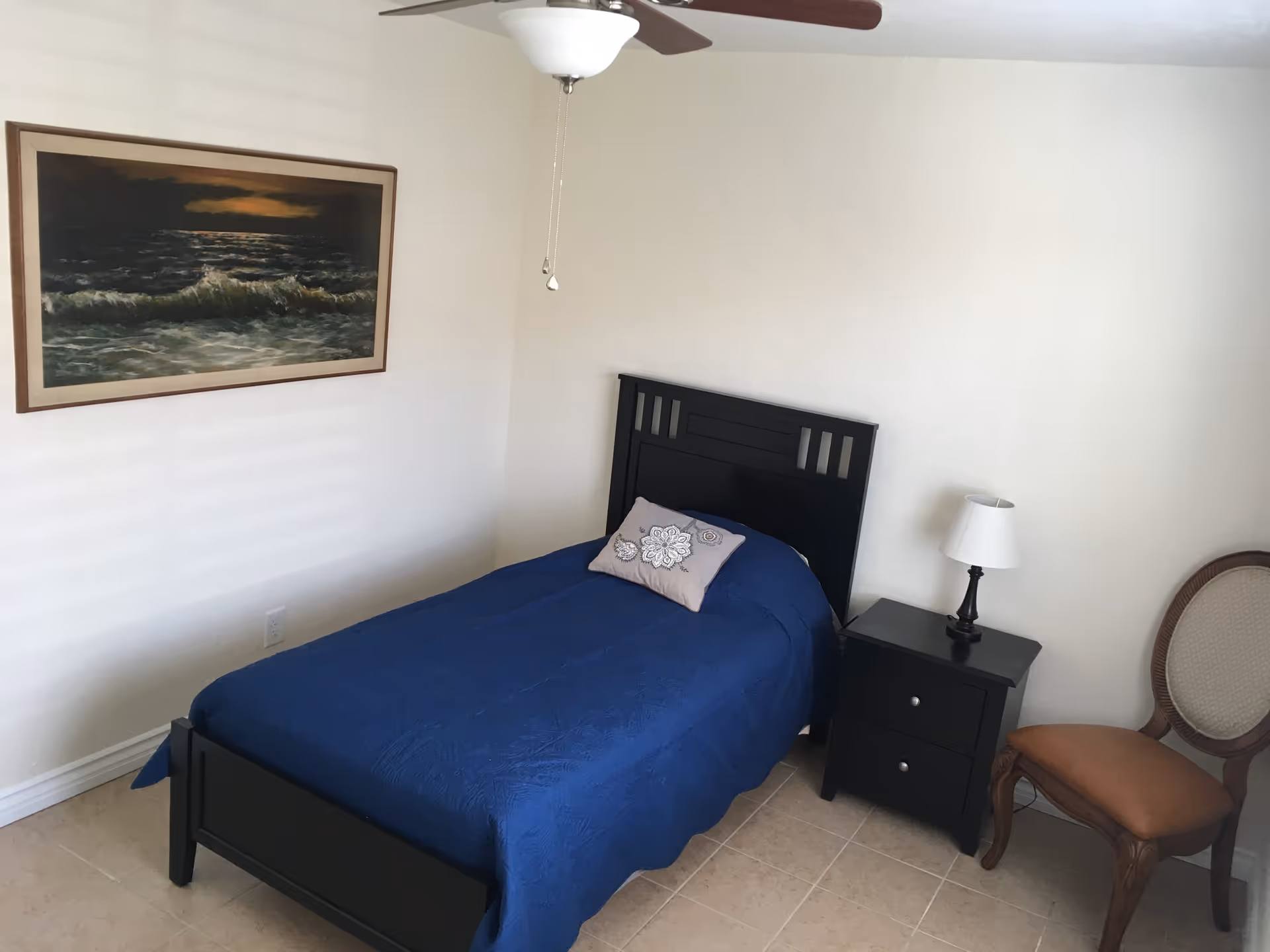 A simple bedroom with a single black bed frame covered with a blue bedspread and a decorative pillow. Next to the bed is a black nightstand with a small lamp. A wooden chair with a cushioned seat is placed beside the nightstand. A ceiling fan with a light fixture hangs above, and a framed painting of ocean waves is mounted on the wall.