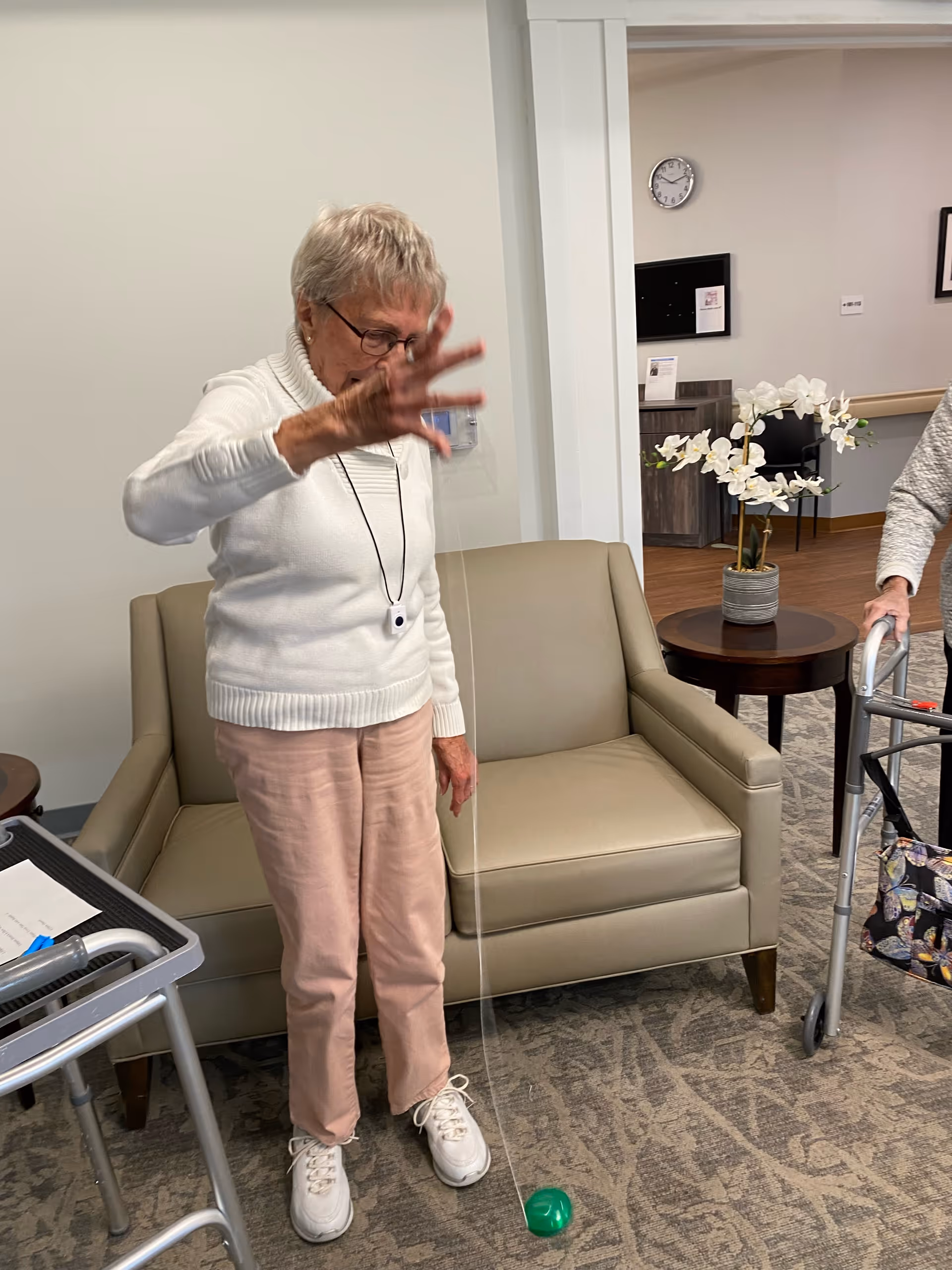 An older woman in a white sweater swings a green ball on a string while standing in a senior living facility lounge near a sofa and walker.