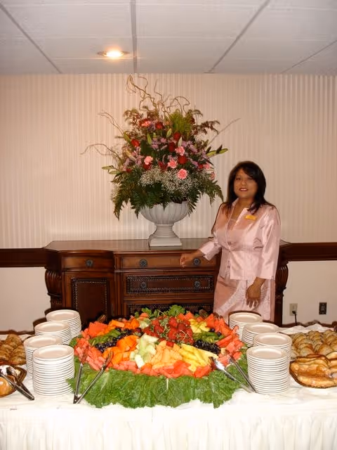 A buffet table with a large fruit platter, stacks of plates and pastries in front of a floral centerpiece and a woman standing beside it.