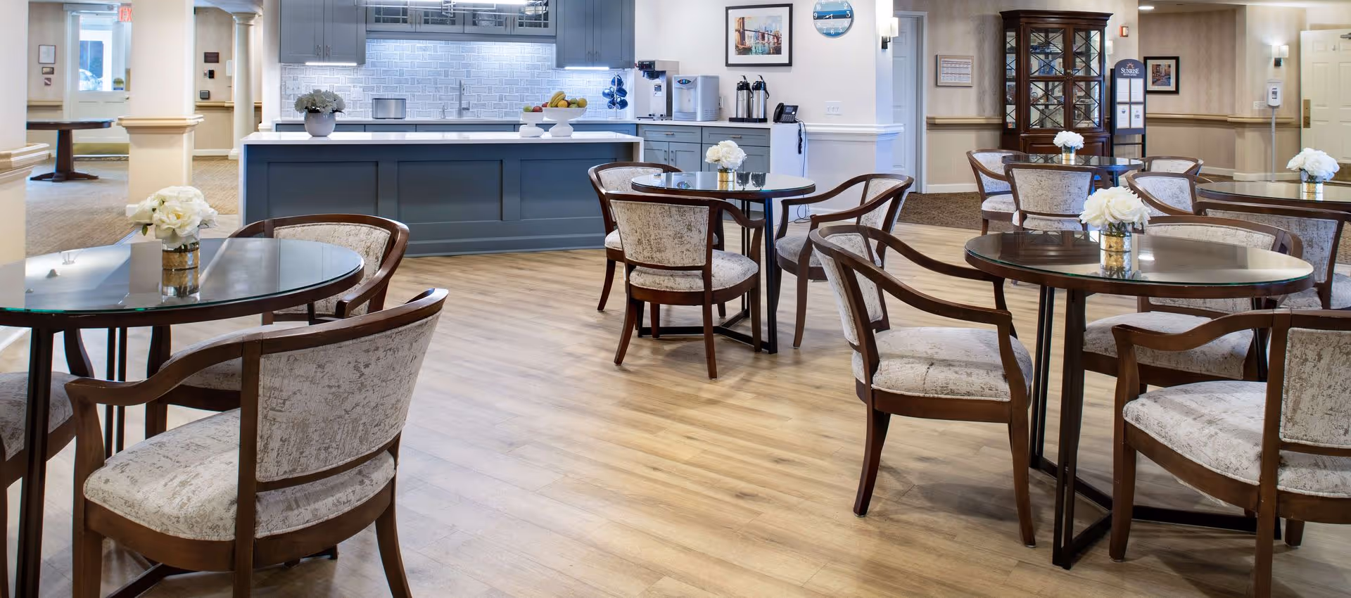 Bright communal dining area with round glass-top tables and upholstered chairs in front of a kitchenette/serving counter.
