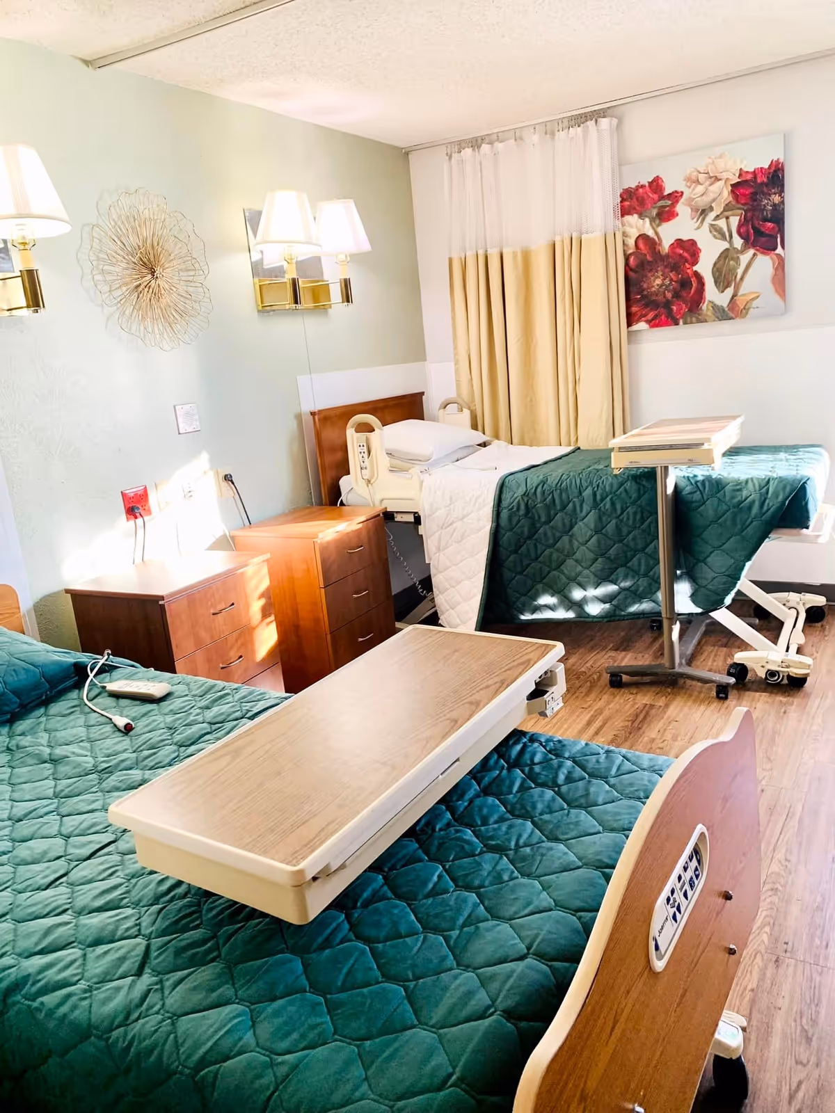 Two adjustable beds with green quilts, overbed tables, bedside drawers, wall lamps and floral artwork in a bright senior care bedroom.
