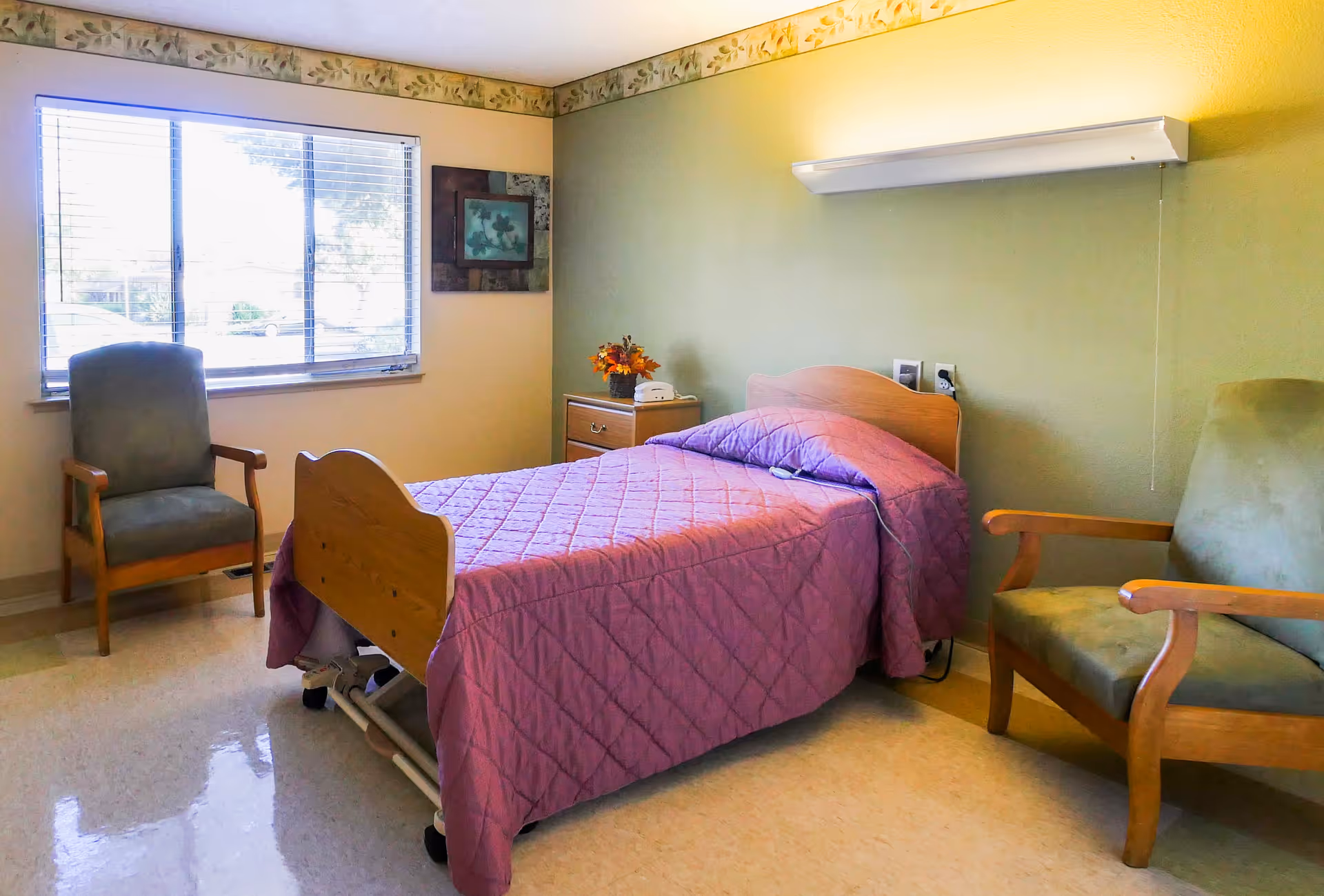 A simple, clean bedroom in a skilled nursing facility with a single bed covered in a purple quilt, two wooden armchairs with green cushions, a small wooden nightstand with a telephone and a flower arrangement, a large window with blinds, and a framed picture on the wall.