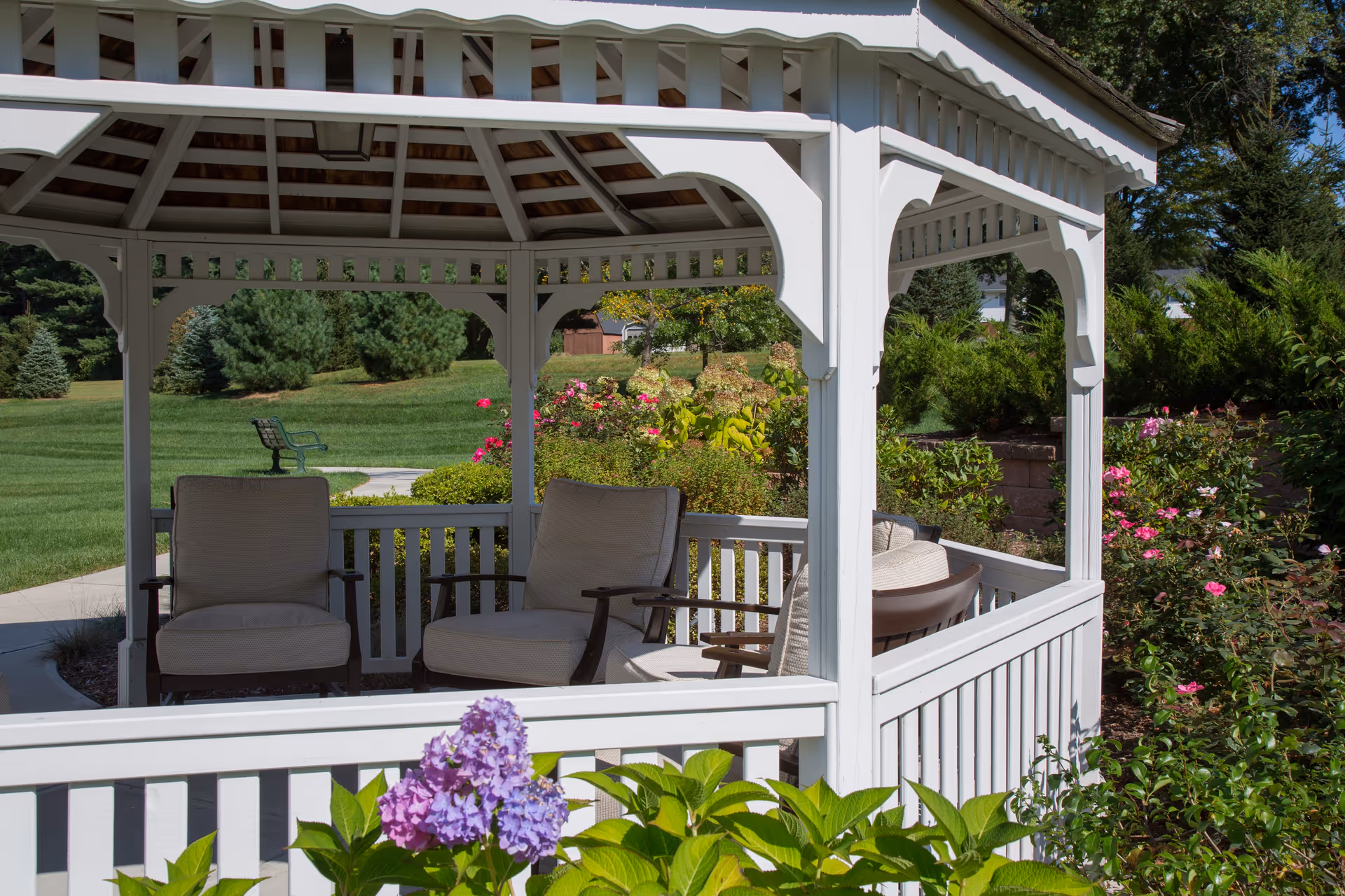 A white wooden gazebo with cushioned chairs inside, surrounded by green grass, bushes, and blooming flowers in a garden setting on a sunny day.
