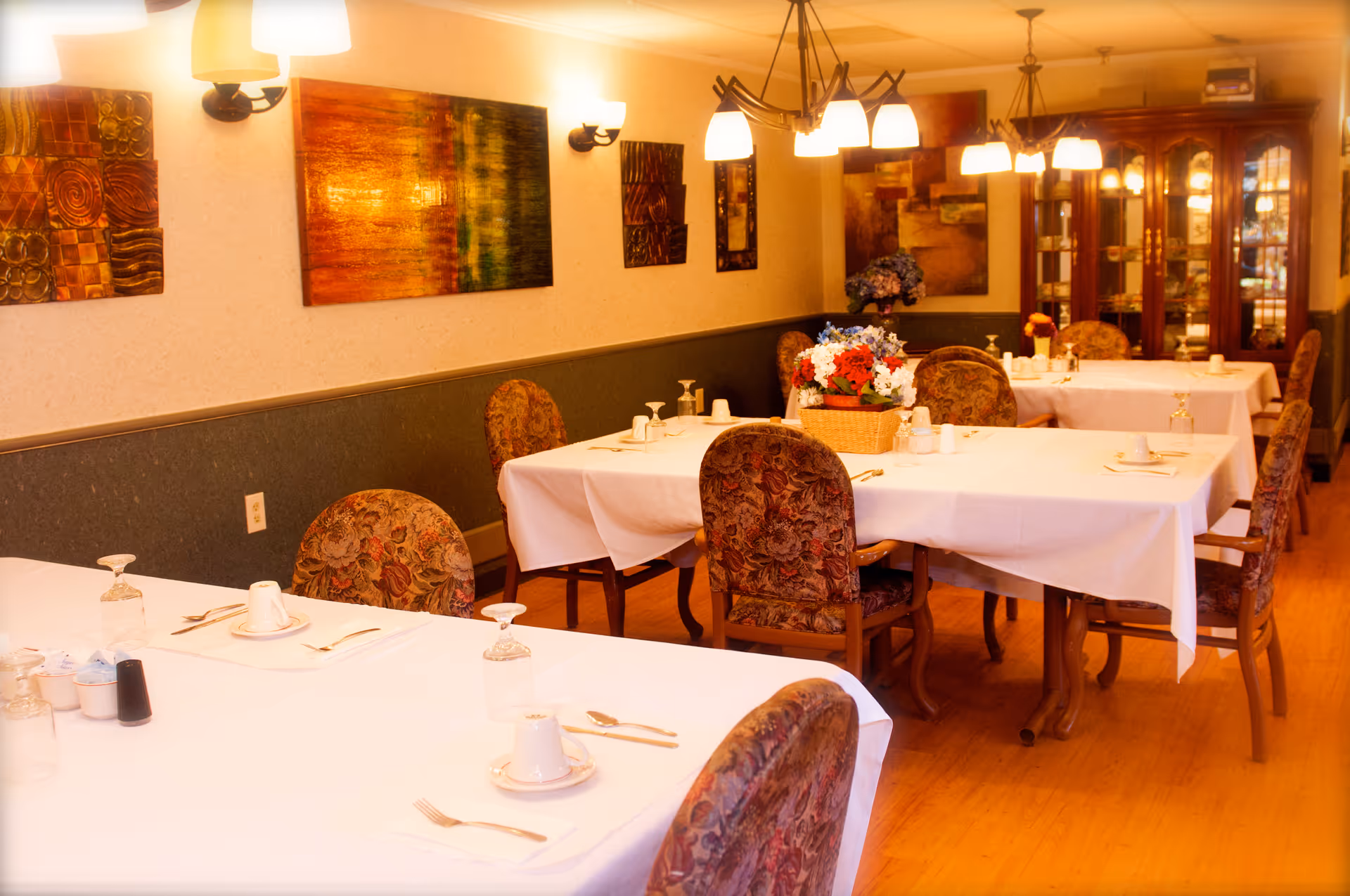 A warmly lit dining room with several tables covered in white tablecloths, each set with cups, saucers, utensils, and upside-down glasses. The chairs have floral upholstery, and there are decorative paintings on the walls. A basket with colorful flowers is placed on one of the tables, and a wooden cabinet with glass doors is visible in the background.