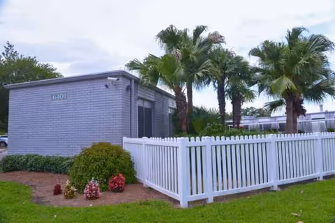 Small gray facility building with a white picket fence and palm trees in front yard.