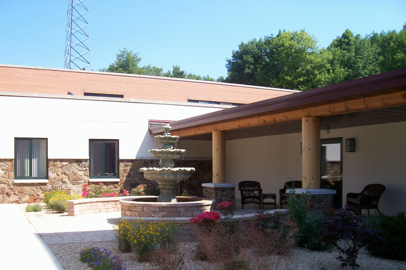 Outdoor courtyard area of Northland Lodge Assisted Living featuring a three-tiered stone fountain surrounded by flower beds and shrubs. There is a covered patio with wooden pillars and several wicker chairs arranged for seating. The building exterior has stone and brick walls with windows and a door leading to the patio.