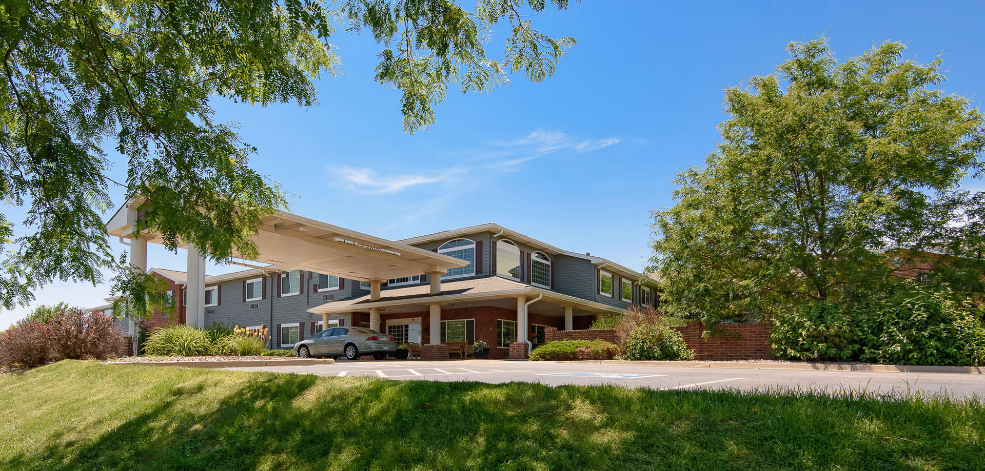 Exterior view of a two-story senior living facility building with a covered entrance, surrounded by green trees and bushes under a clear blue sky.
