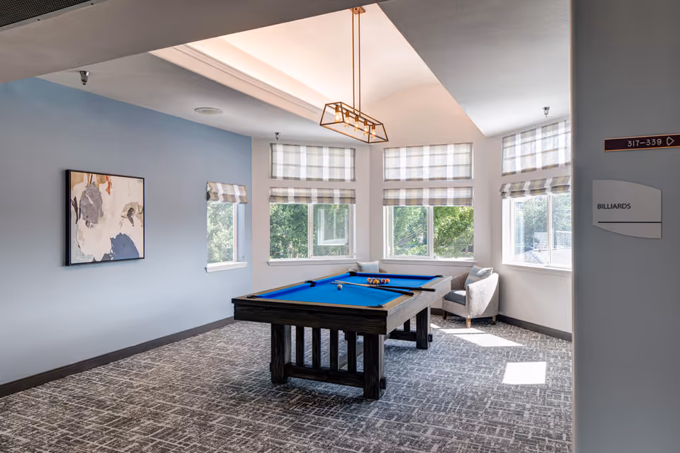A bright room with large windows featuring checkered blinds, a blue felt billiards table in the center, a modern light fixture hanging from the ceiling, a gray armchair in the corner, and a piece of abstract art on a light blue wall.