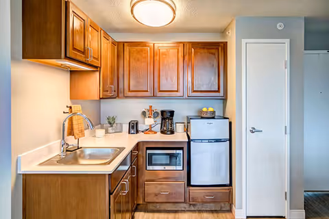 Small kitchenette with wooden cabinets, a stainless sink, microwave, mini refrigerator, and countertop appliances.