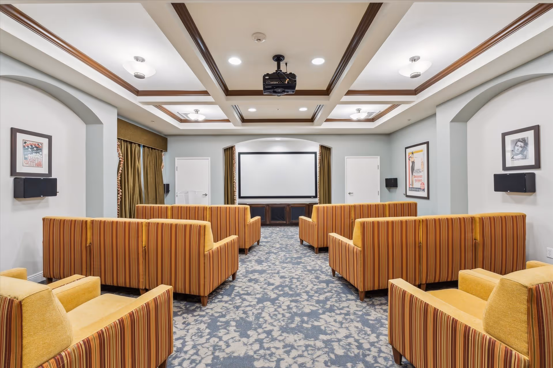 A cozy media room with multiple striped orange and yellow armchairs arranged in rows facing a large projection screen. The room has a patterned carpet, framed artwork on the walls, and a ceiling-mounted projector with recessed lighting.