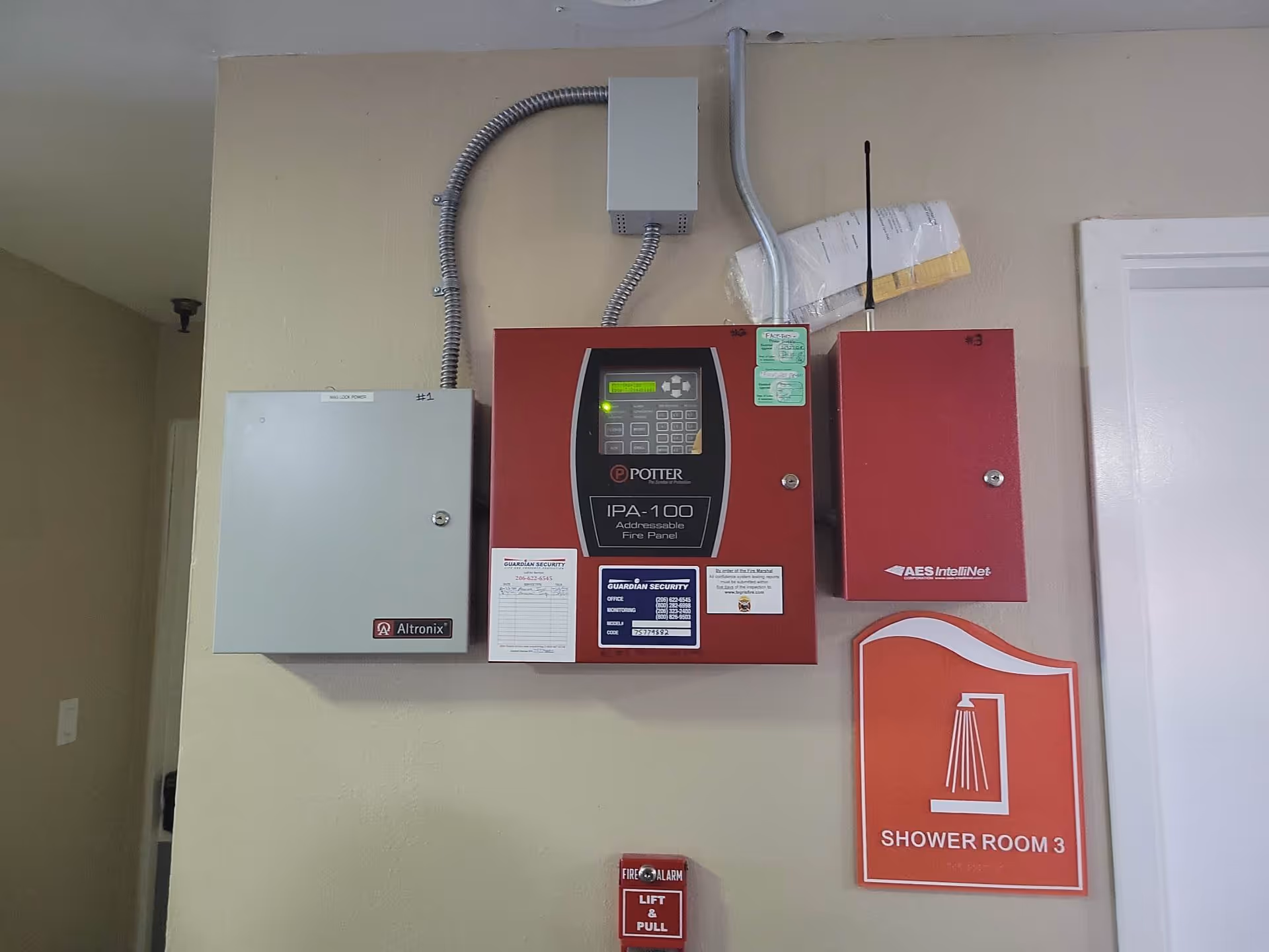 Wall-mounted fire alarm control panels and electrical boxes with a red sign below indicating Shower Room 3, and a fire alarm pull station beneath the sign.