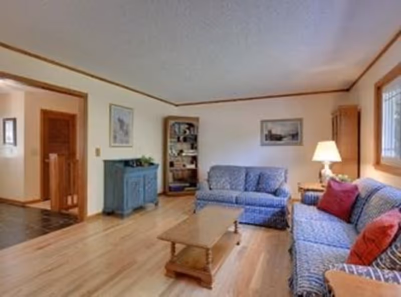 A cozy living room with wooden flooring, two blue patterned sofas with red cushions, a wooden coffee table in the center, a blue cabinet against the wall, a tall bookshelf in the corner, a table lamp on a side table, and framed artwork on the walls. There is a window letting in natural light on the right side.