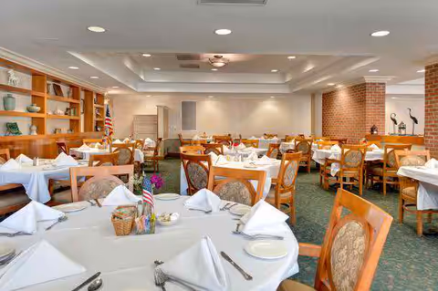 Bright dining room with multiple tables set with white tablecloths, napkins and place settings, wooden chairs, shelving and an American flag on one table.