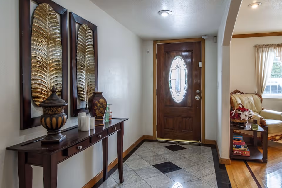 Entryway of a senior living facility with a wooden front door featuring an oval glass panel. To the left, a dark wooden console table holds decorative items including a vase, candles, and a hand sanitizer bottle. Above the table are two framed metallic leaf wall decorations. To the right, an archway leads to a living room area with a cream-colored armchair, a side table with a basket of flowers, and a window with curtains.