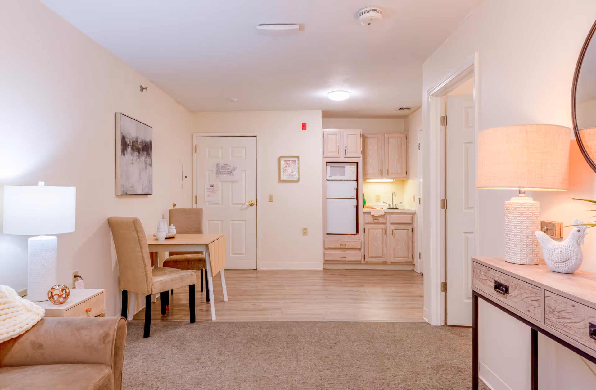 A bright furnished living area with a small dining table, kitchenette, and lamps.