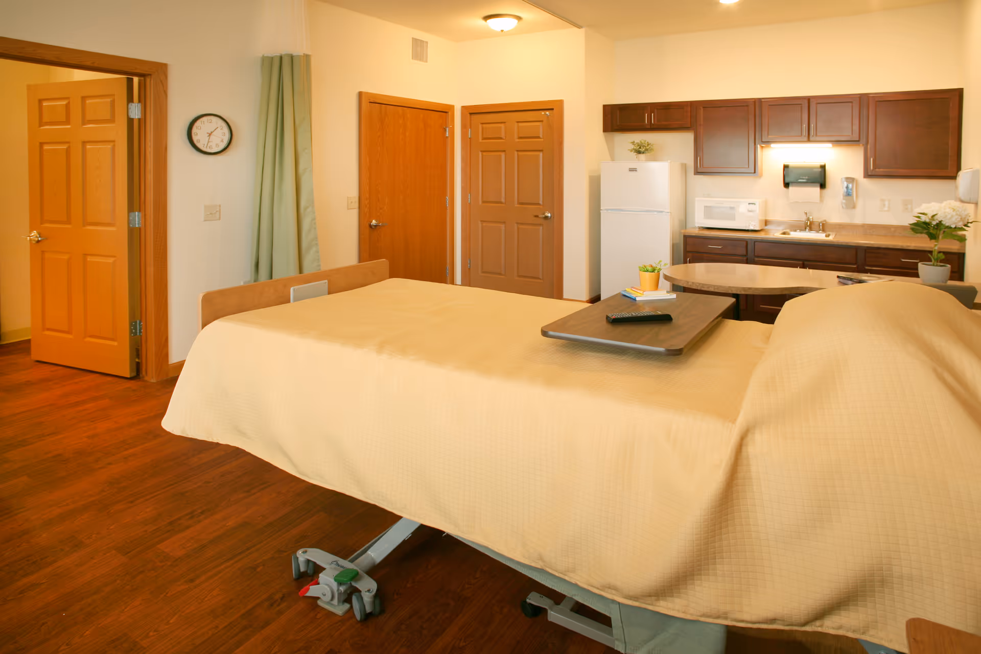 A senior living facility room with a hospital-style bed covered in a beige blanket, a wooden tray with a remote control on the bed, wooden doors, a clock on the wall, and a kitchenette area with a refrigerator, microwave, sink, and cabinets.