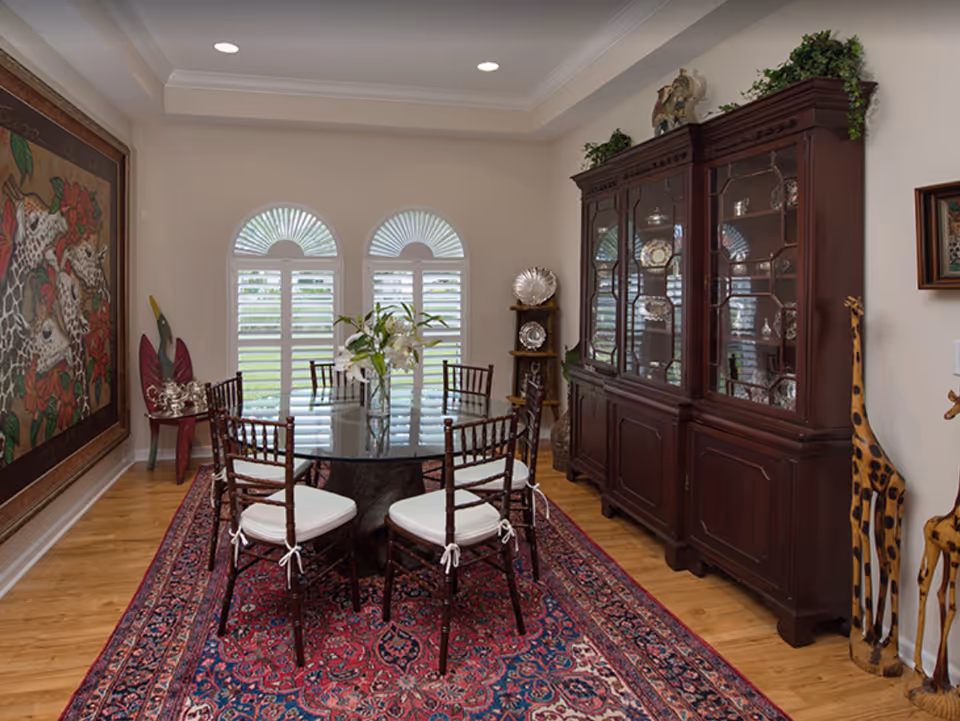 A dining room with a round glass table surrounded by six wooden chairs with white cushions. The floor has a large red and blue patterned rug. Against the wall is a dark wooden china cabinet displaying silver plates and decorative items. There are three arched windows with white shutters letting in natural light. The room also features a large painting of giraffes on the left wall and two tall wooden giraffe sculptures on the right side.
