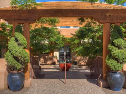 Wooden arched entrance with potted evergreens leads to a brick senior living facility exterior.