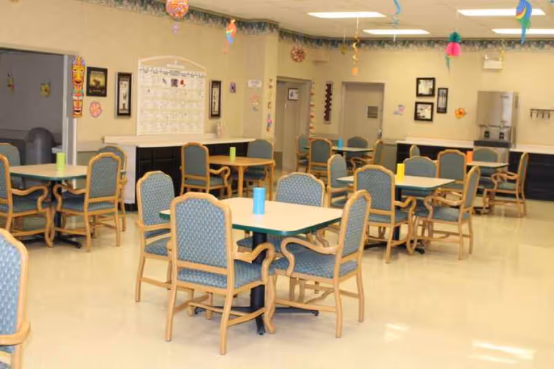 A dining room in a senior living facility with multiple tables and chairs arranged neatly. The room is decorated with colorful hanging ornaments and has a bright, clean appearance. There is a calendar and framed pictures on the walls, and a counter area with a coffee machine in the background.