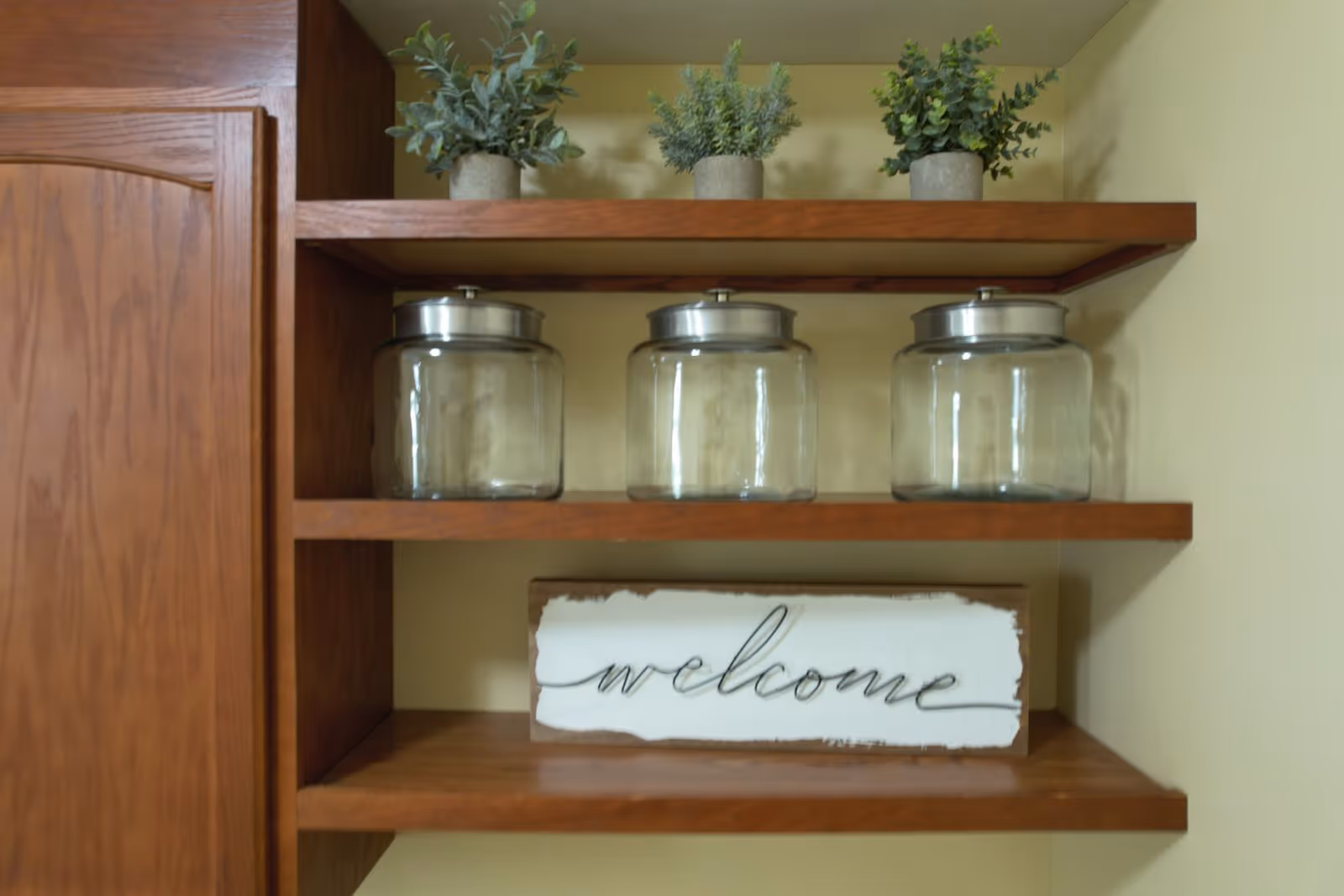 Wooden shelves mounted on a beige wall with three small potted plants on the top shelf, three empty glass jars with metal lids on the middle shelf, and a decorative wooden sign with the word 'welcome' on the bottom shelf.