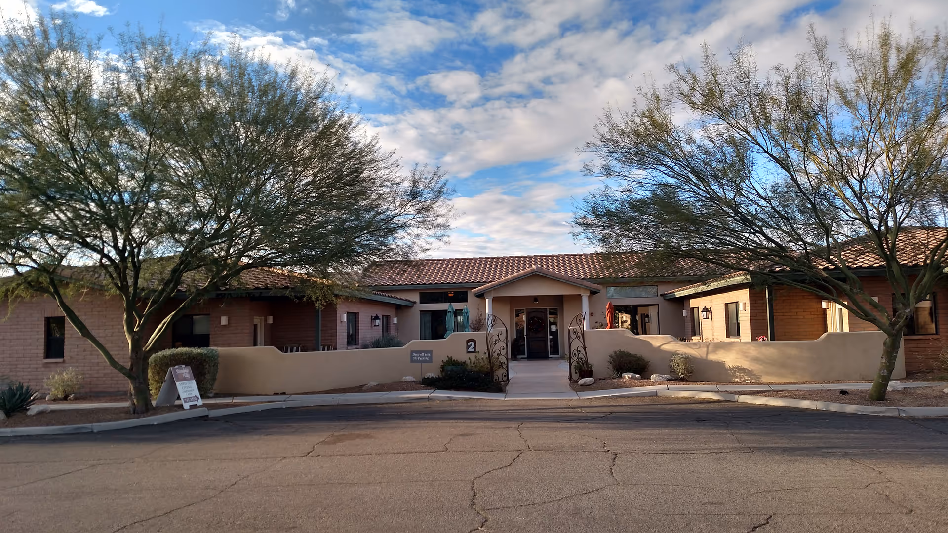 Front exterior view of BridgeWater Assisted Living facility in Tucson, showing a single-story building with a tiled roof, beige walls, and a gated entrance. Two large trees flank the entrance, and there is a sign on the left side near the curb. The sky is partly cloudy with blue patches.
