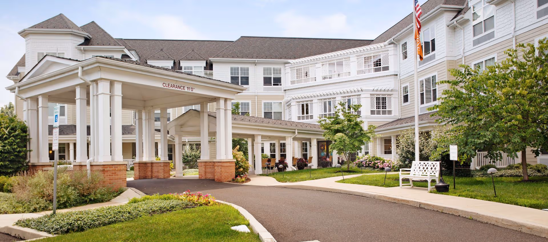 Exterior view of Sunrise of North Wales senior living facility showing a covered entrance with white columns and brick bases, a driveway, landscaped greenery, trees, a white bench, and an American flag on a flagpole.