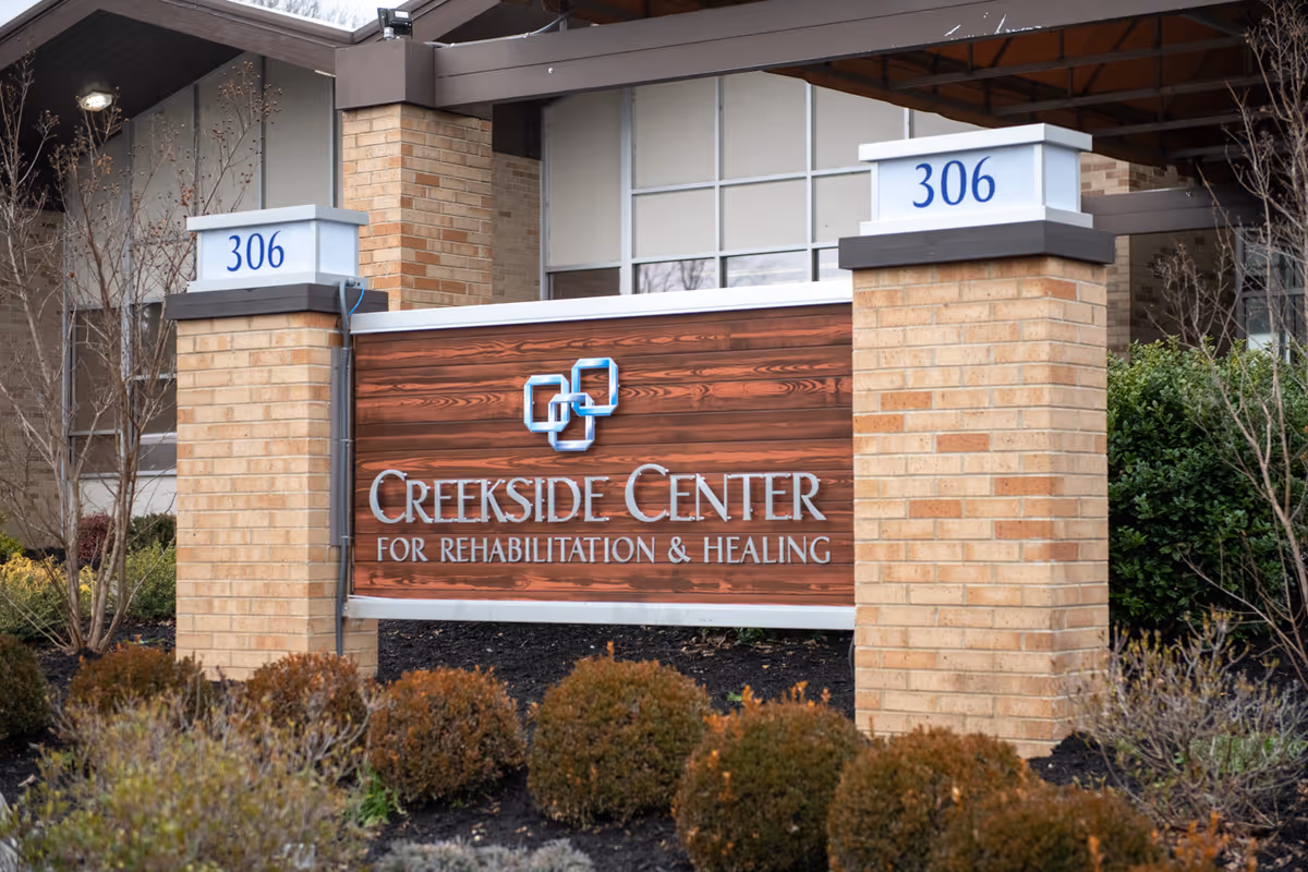 Outdoor view of the entrance sign for Creekside Center for Rehabilitation & Healing, mounted on a wooden panel between two brick pillars with the number 306 on top of each pillar. The background shows part of the building and some landscaping with bushes and trees.