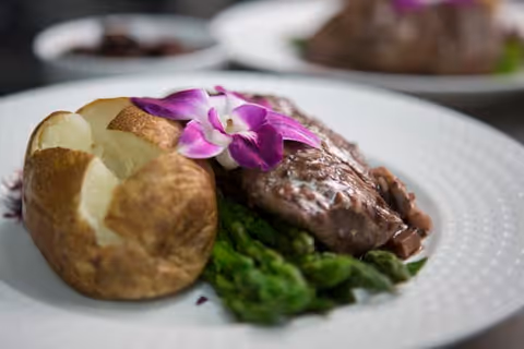 A plated meal featuring a baked potato, a serving of asparagus, and a piece of meat topped with a purple orchid flower as garnish.
