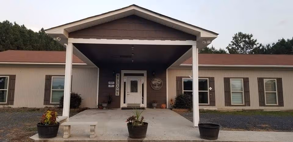 Front exterior view of Autumn Estate Personal Care Home showing a single-story building with a covered entrance supported by white pillars, a concrete walkway, potted plants, and a welcome sign near the door.