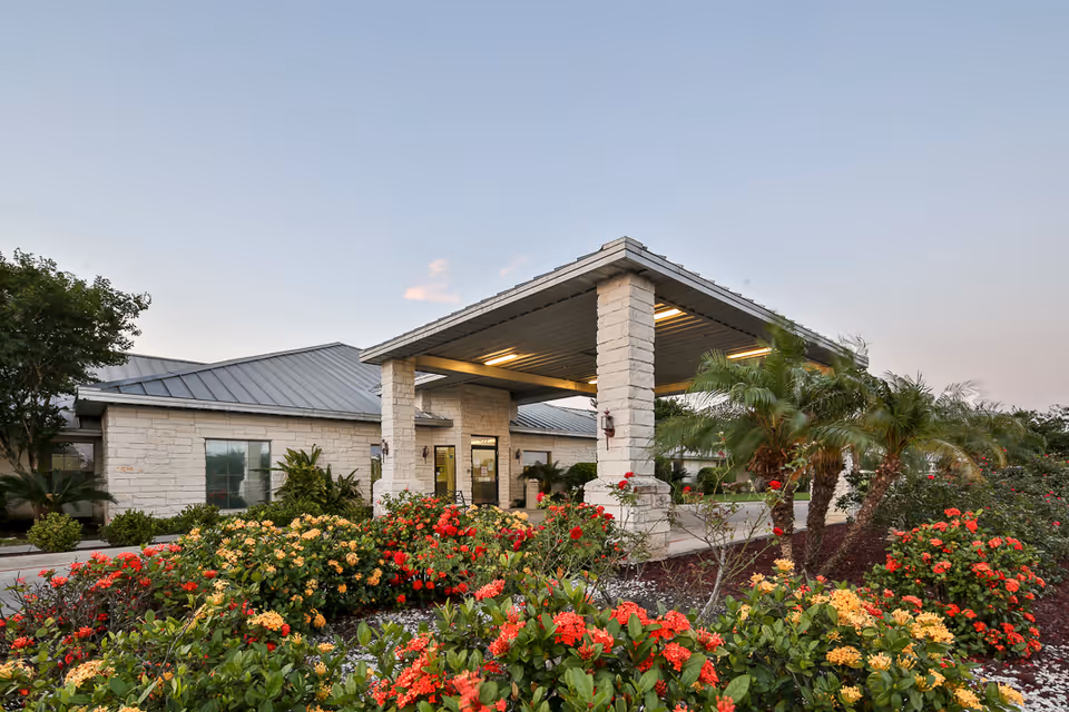 Covered entrance and drop-off canopy of a single-story stone-clad nursing facility surrounded by landscaped flower beds and palm trees.