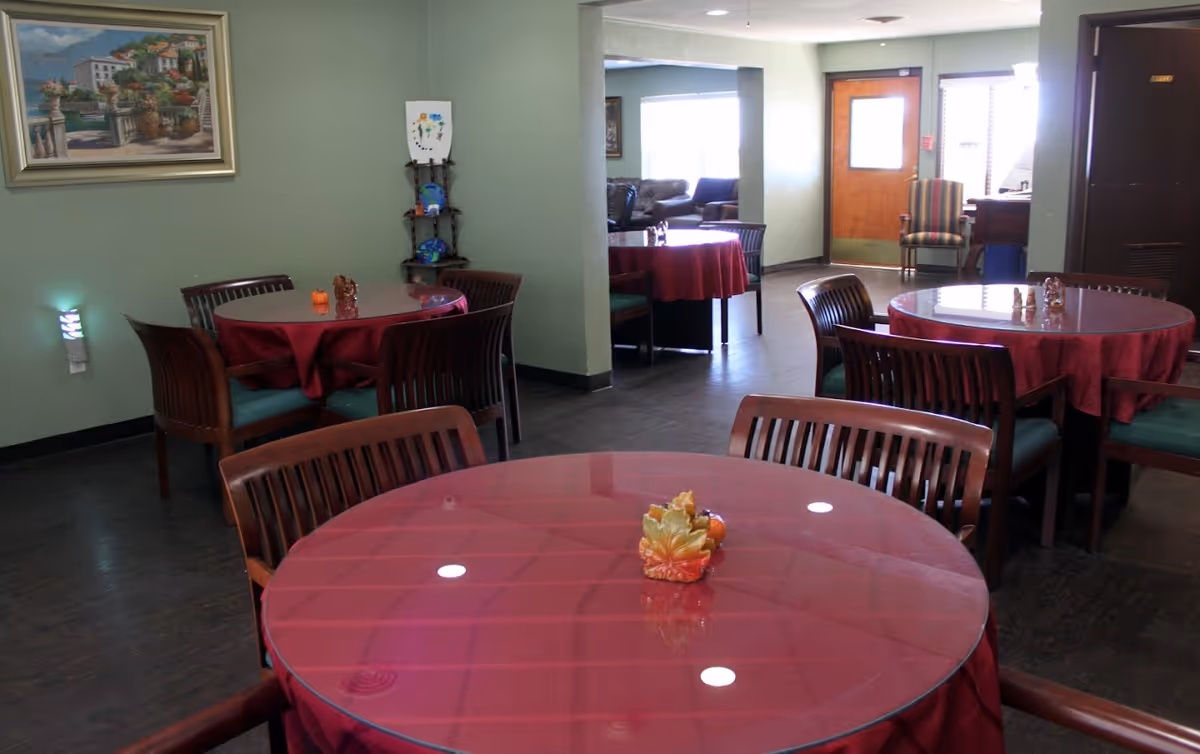 Interior view of a dining area in Silver Creek Assisted Living Azle with round tables covered in red tablecloths and wooden chairs around them. The room has light green walls, a painting on one wall, a small decorative shelf, and a door leading to another room with seating visible.