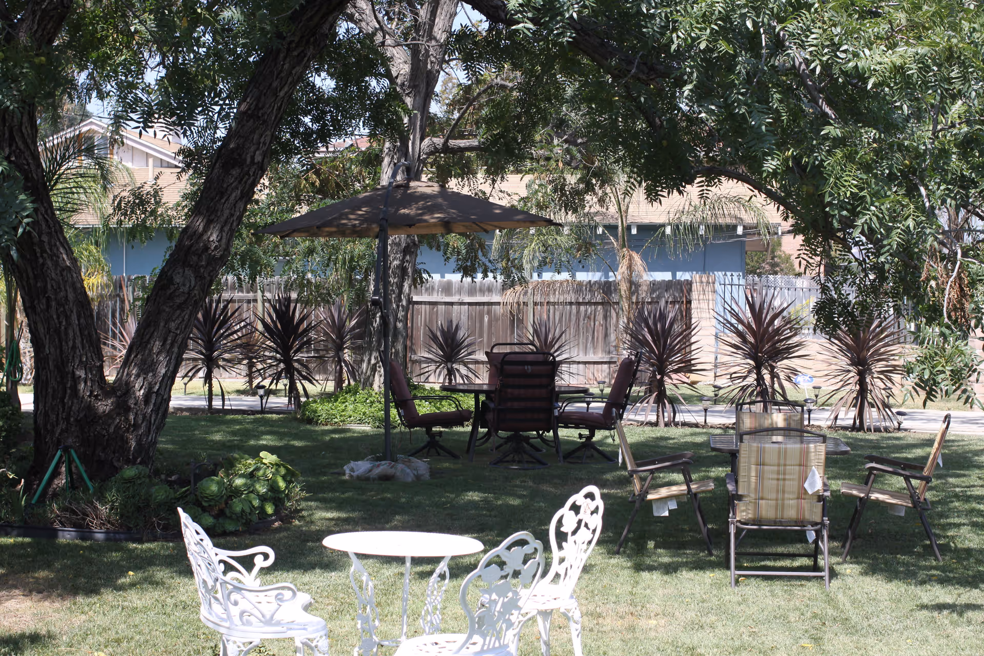 Outdoor garden area with green grass, several trees providing shade, and multiple seating arrangements including white metal chairs with a small round table and cushioned chairs around a table with an umbrella. A wooden fence and neighboring houses are visible in the background.